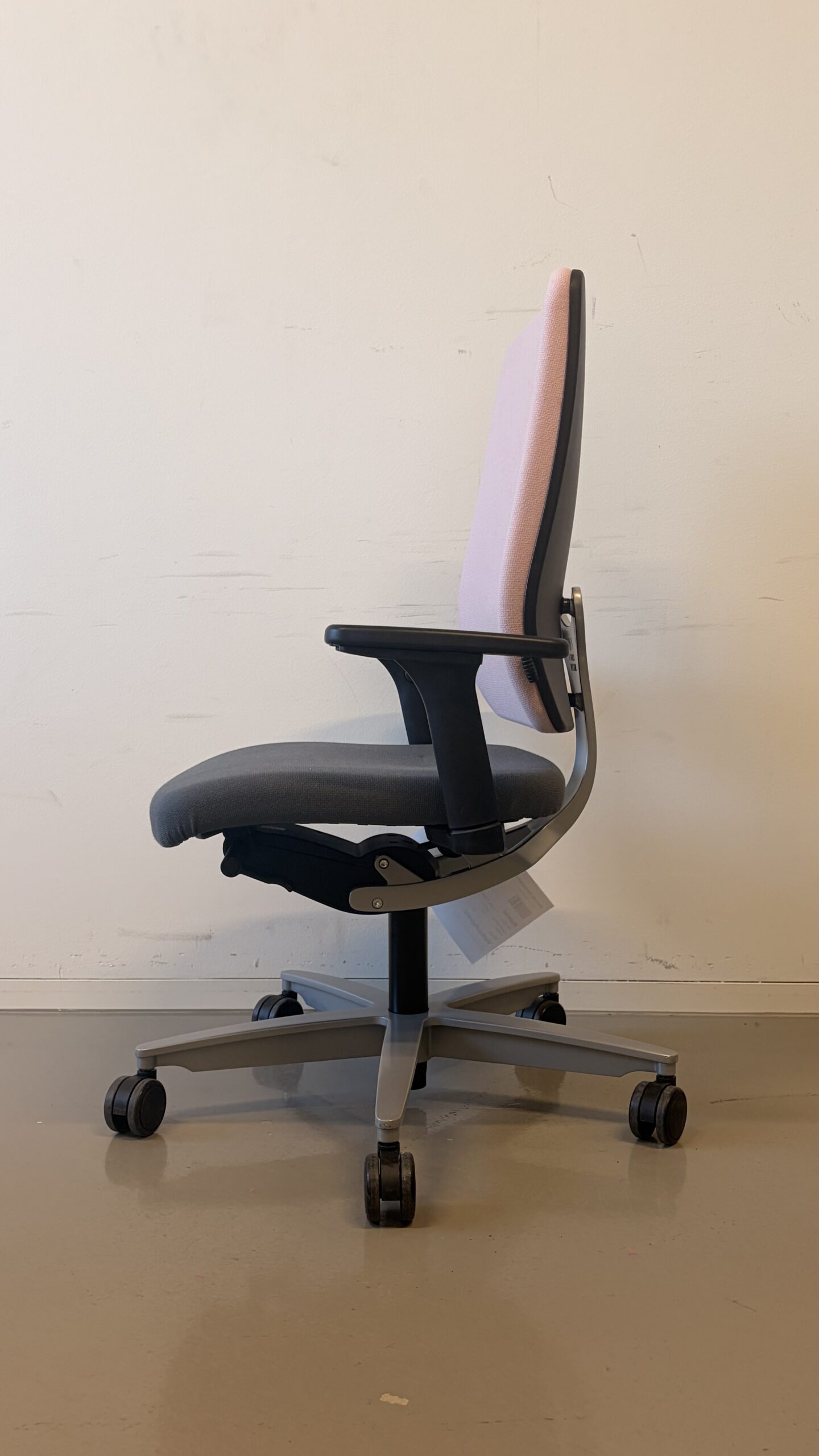 Bürostuhl mit hellrosa Rückenlehne, grauem Sitz und schwarzen Armlehnen vor weißer Wand seitlich